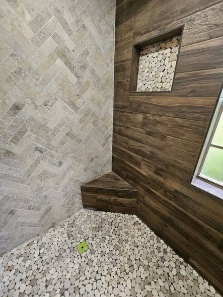 A custom tile shower by Level Headed Construction with herringbone stone-look walls, a wood-look plank accent wall, a pebble floor, and a built-in bench