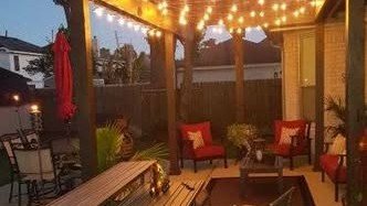 A covered backyard patio with a pergola-style cover strung with lights over red outdoor furniture at dusk in the Huffman / Lake Houston area