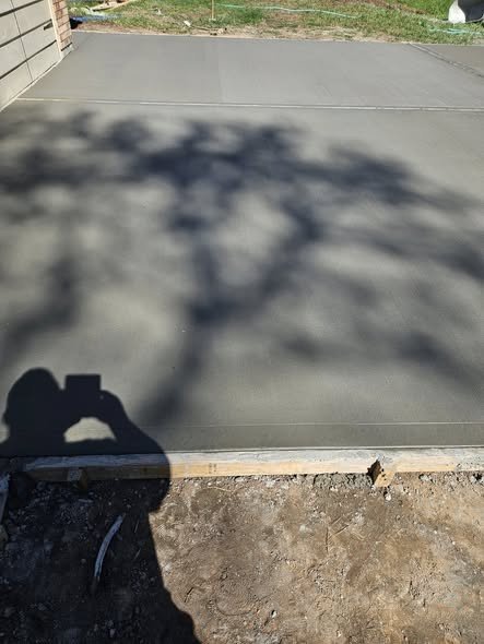 A freshly poured concrete foundation slab curing inside its wood form boards, with dappled tree shadows, in the Huffman / Lake Houston area
